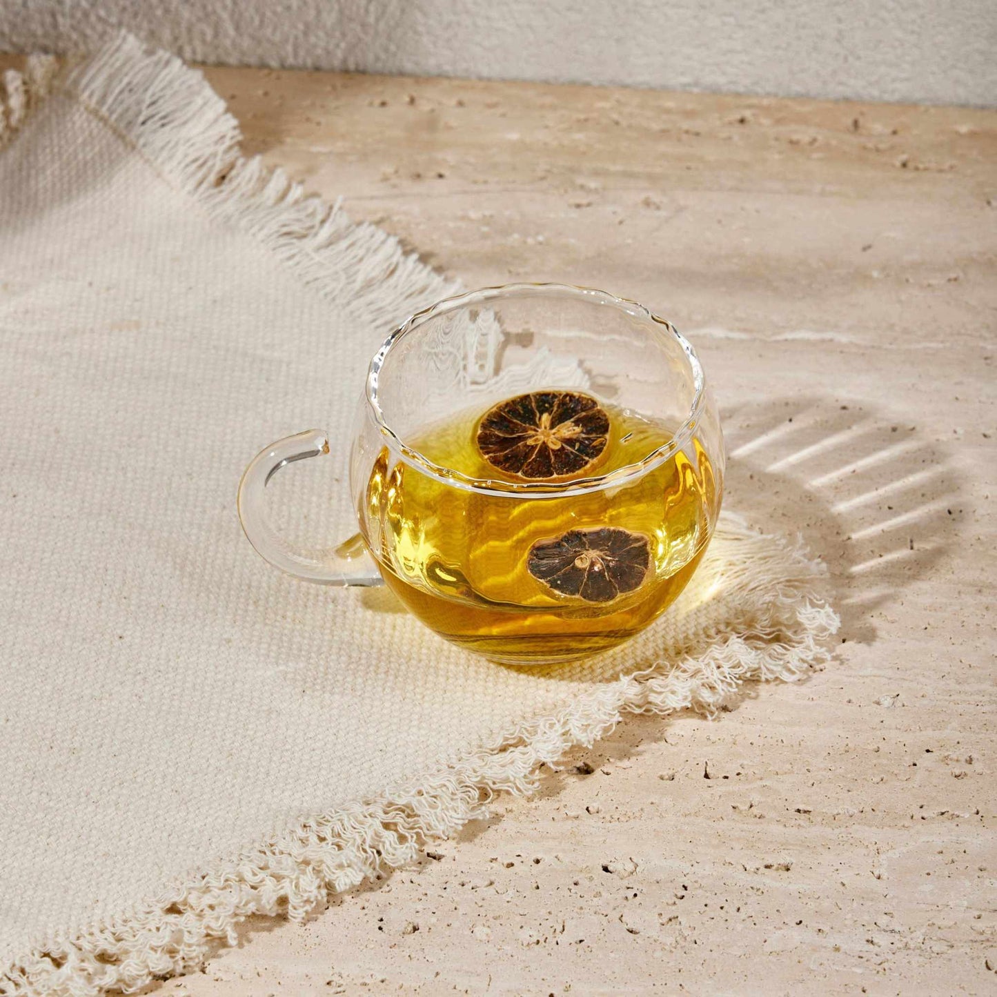 Transparent glass KURE CUP with tea and a slice of dried citrus on a textured surface.