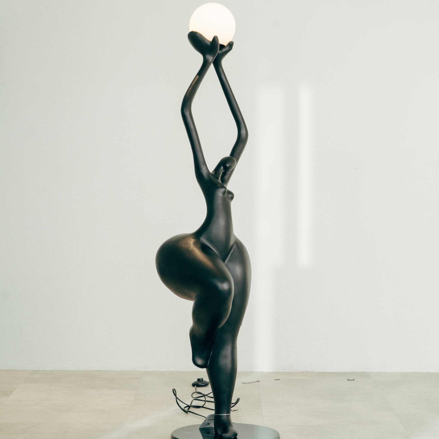 Matte black abstract human form floor lamp holding luminous globe.