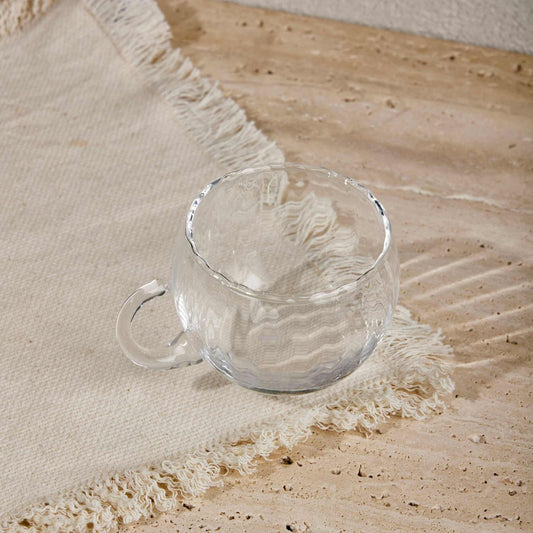 Transparent glass KURE CUP with ergonomic handle on textured fabric background.