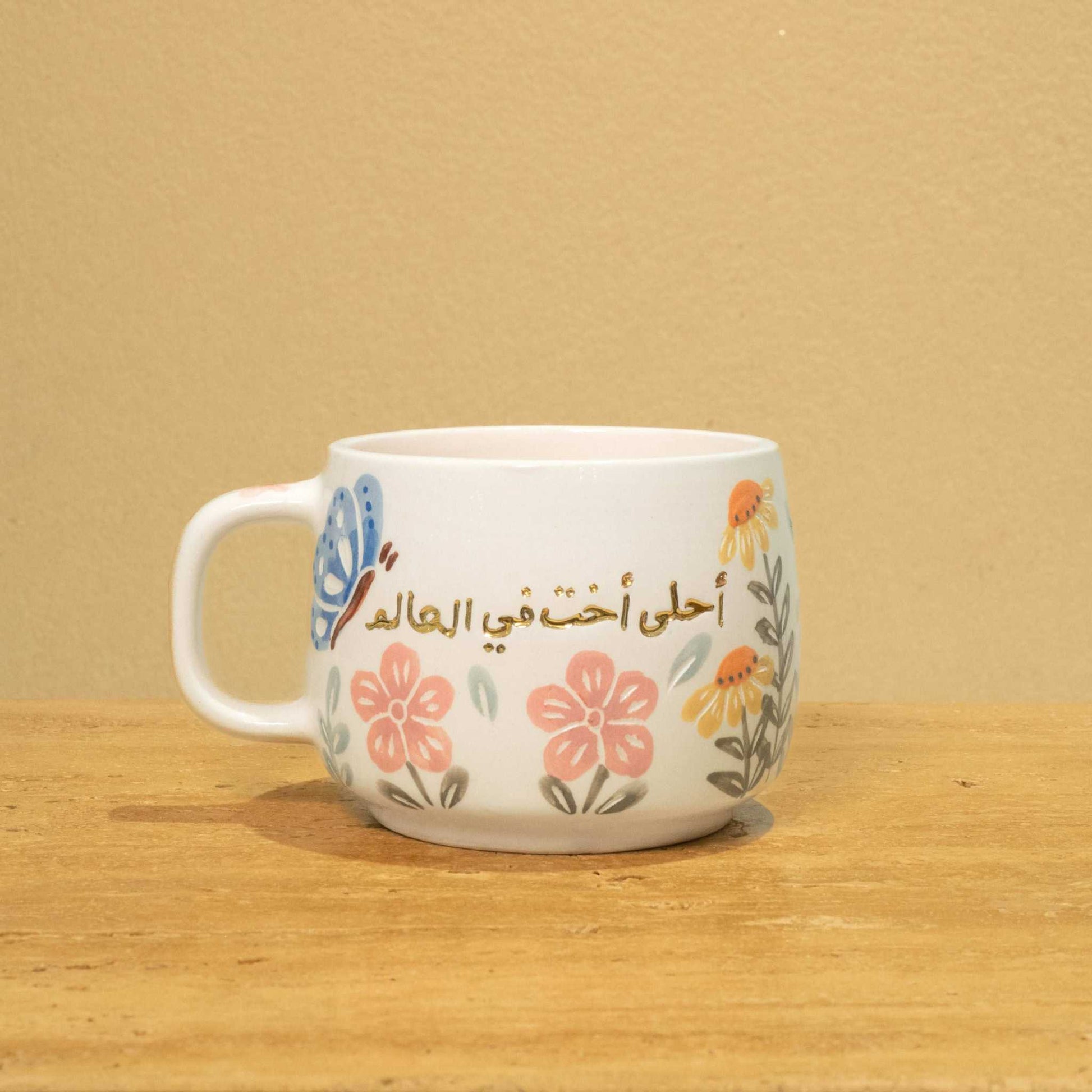 Gold Arabic monogram ceramic mug with floral design, "Best Sister in the World".