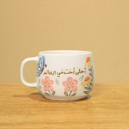 Gold Arabic monogram ceramic mug with floral design, "Best Sister in the World".