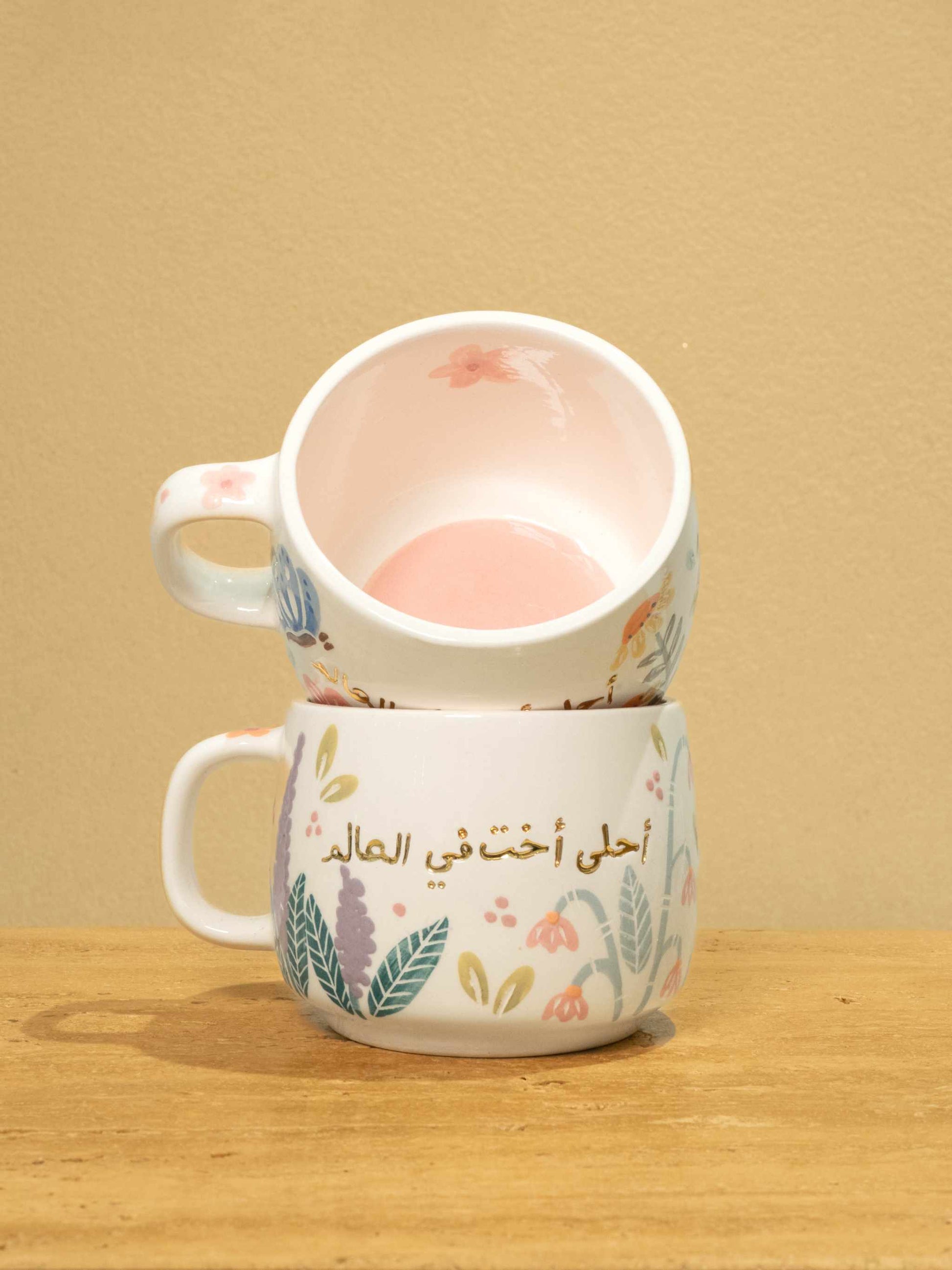 Ceramic mug with gold monogram "أحلى أخت في العالم" and floral design.